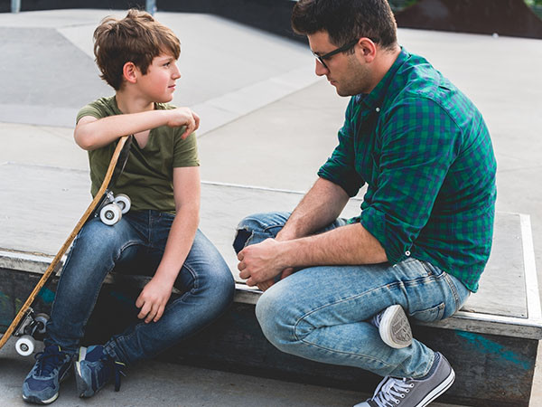 父子面对面坐在滑板公园的混凝土路边交谈，儿子的腿上靠着一块滑板