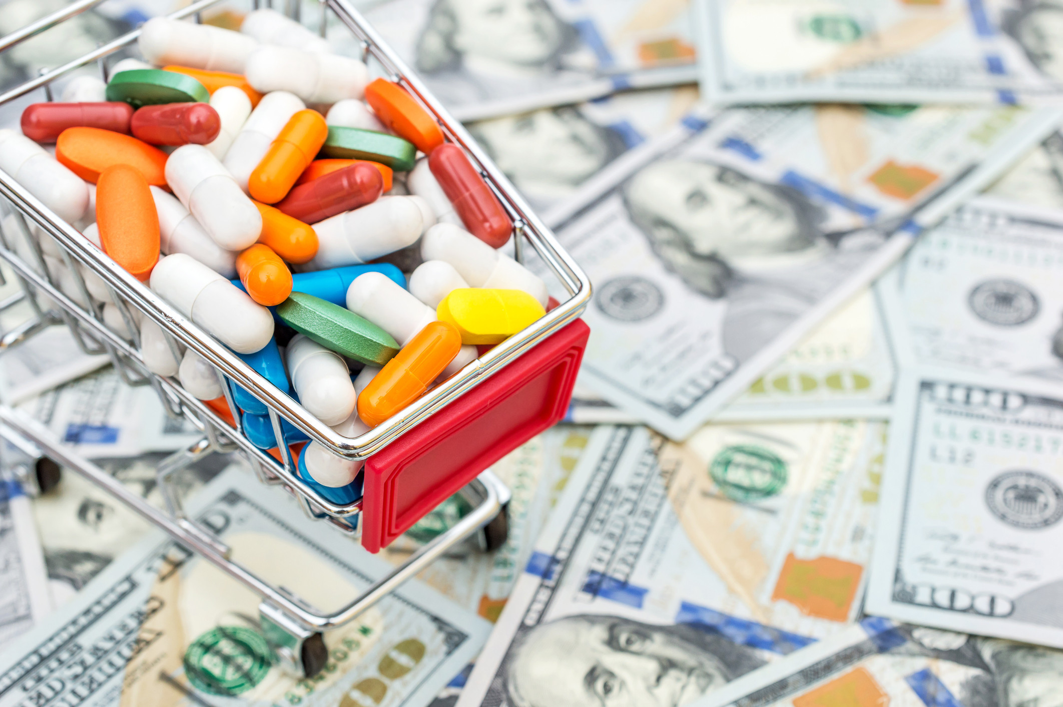 A tiny shopping cart filled with colorful pills and tablets is parked on top of hundred dollar bills