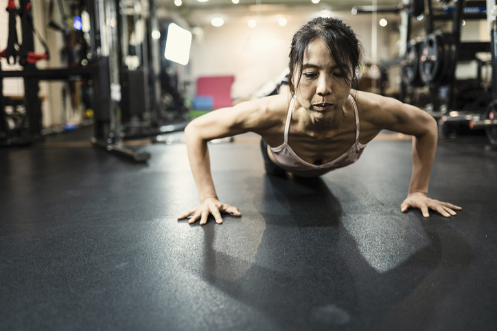 A mature Asian woman in plank position on the floor of a gym.
