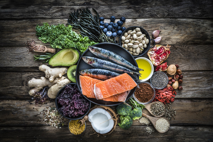 overhead view photo of a selection of healthy foods arranged on a wooden table, including fruits and vegetables, nuts and grains, fish and olive oil