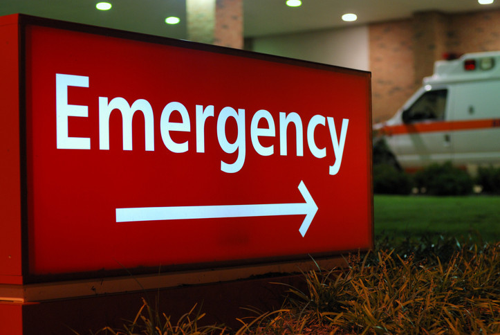 ป้ายแดงขาวนอกโรงพยาบาลที่มีคำว่า "ฉุกเฉิน" ชี้ไปที่แผนกฉุกเฉิน ตอนกลางคืนและมีรถพยาบาลจอดอยู่ข้างนอก