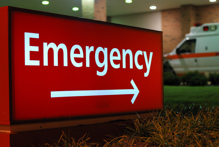 Panneau rouge et blanc à l'extérieur d'un hôpital avec le mot "Urgence" pointant vers le service des urgences ;  il fait nuit et une ambulance est garée à l'extérieur 