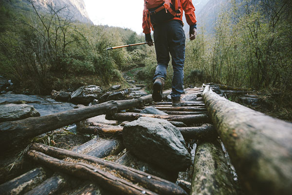 photo taken from behind and close to the ground of a hiker crossing a bridge over a stream made of logs