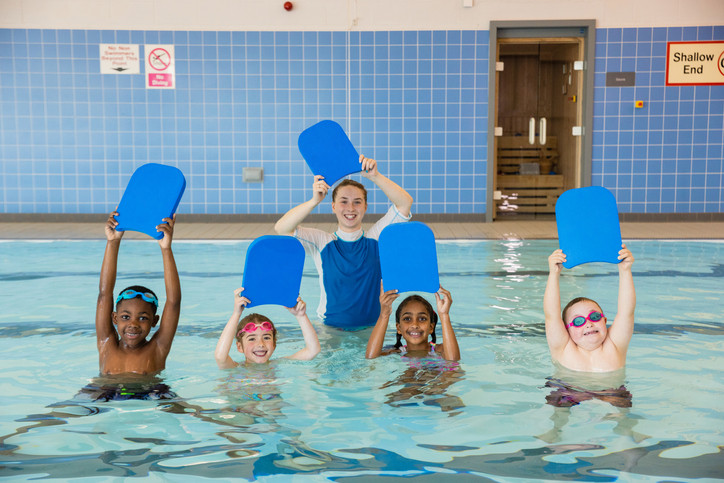 เด็กสี่คนในสระตื้นๆ กำลังเรียนว่ายน้ำกับอาจารย์ผู้สอน เด็ก ๆ กำลังยืนอยู่ในน้ำโดยถือกระดานเตะสีน้ำเงิน