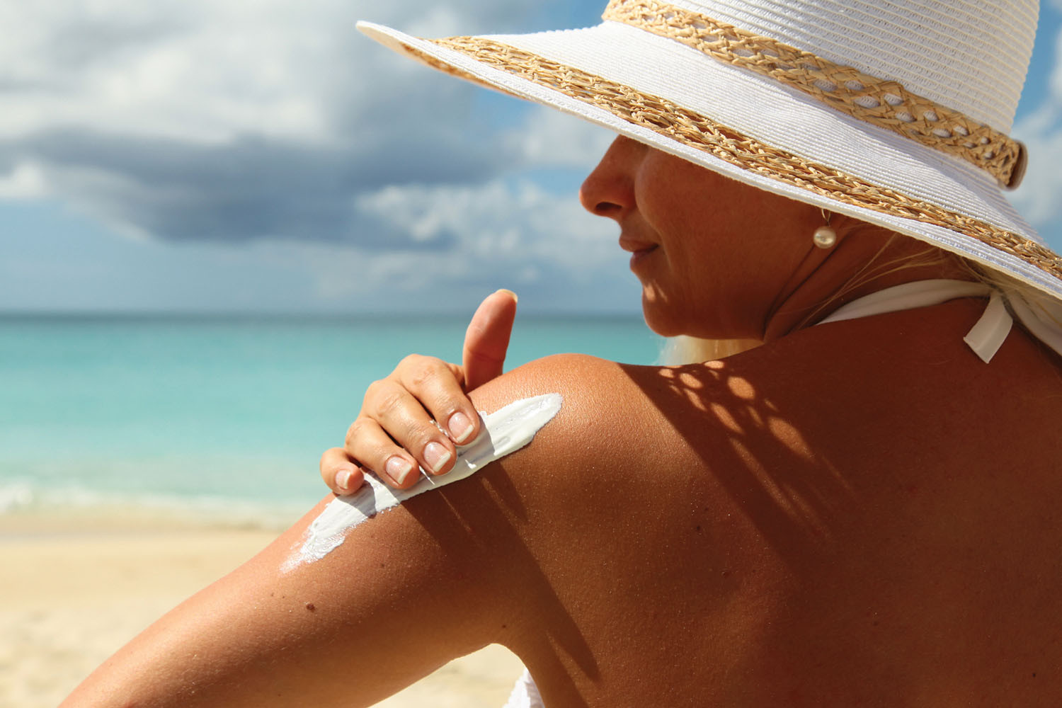 photo of a woman on a beach wearing a sun hat viewed from behind as she applies sunscreen to her upper arm
