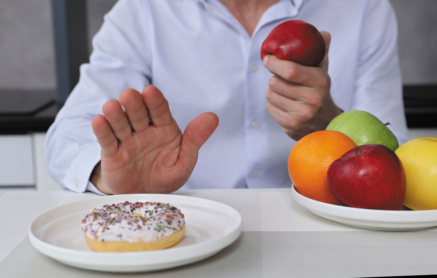 cropped photo showing the hands and torso of a man refusing a doughnut and choosing an apple from a bowl of assorted fruit