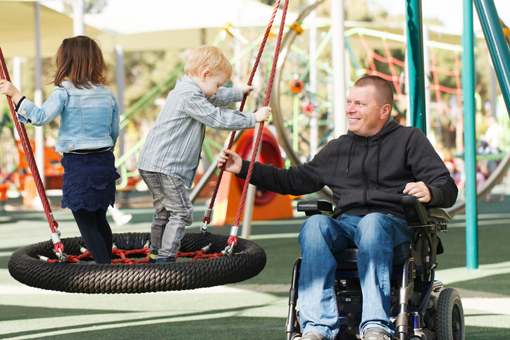 Un père vêtu d'un sweat-shirt foncé et d'un jean est assis dans un fauteuil roulant et joue avec ses deux jeunes enfants sur une balançoire dans une aire de jeux