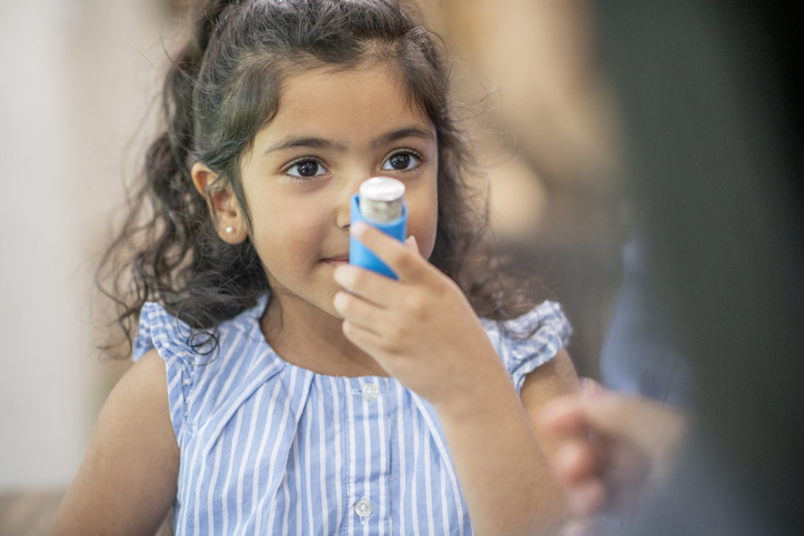 Une enfant aux cheveux et aux yeux foncés portant un haut rayé bleu et blanc apprend à utiliser un inhalateur pour asthmatiques, qu'elle tient près de sa bouche ;  adulte flou vu partiellement de dos