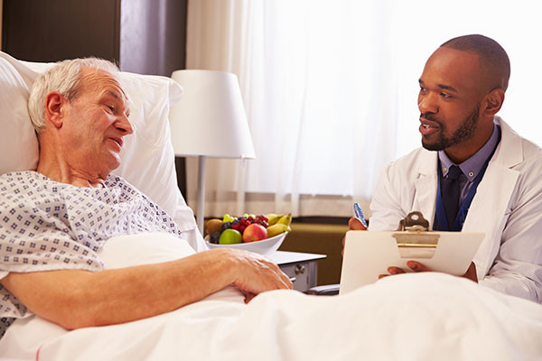 photo of a senior man in a hospital bed talking with a doctor who is sitting next to the bed holding a clipboard