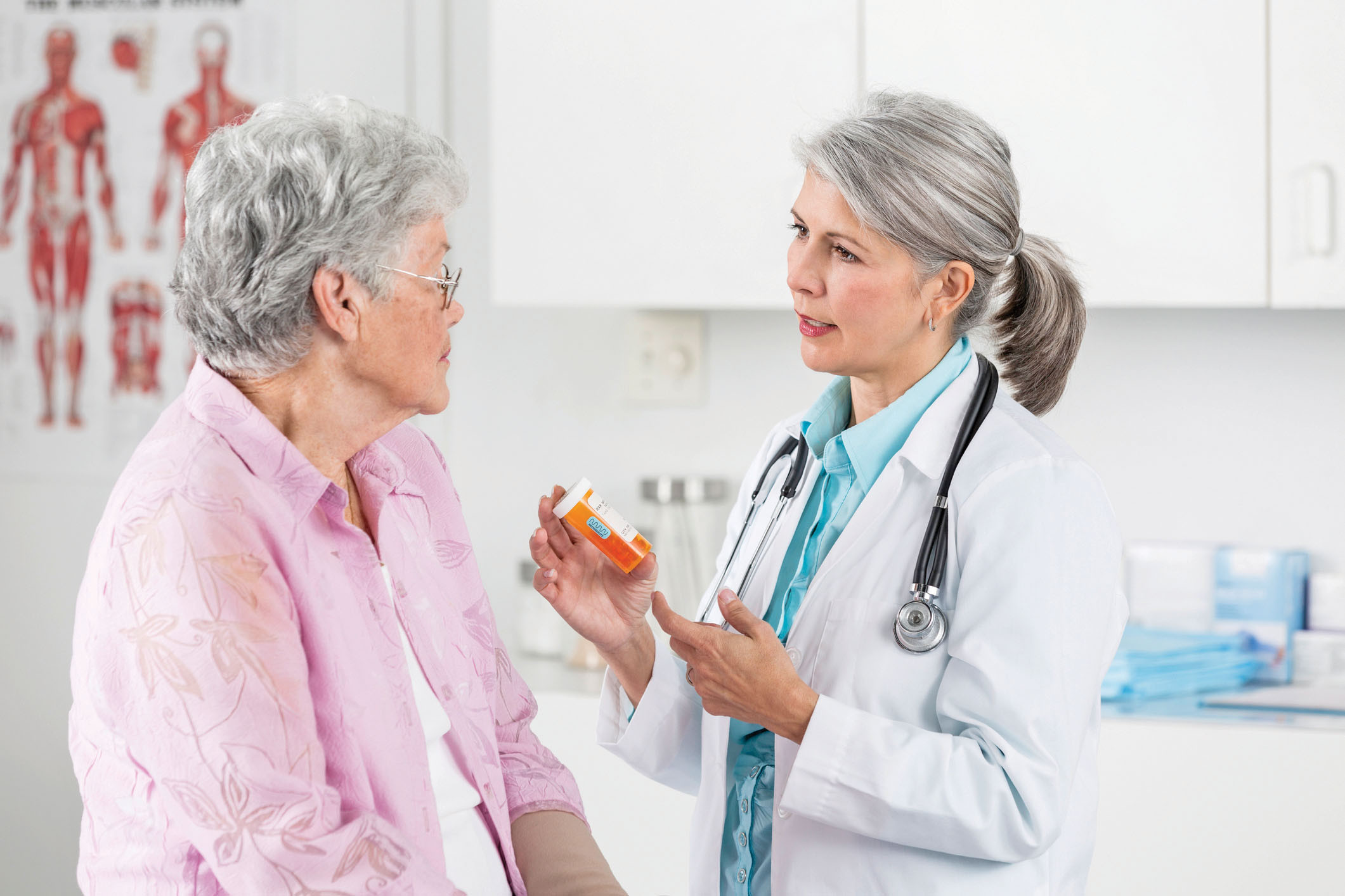 photo of a female doctor speaking with an older female patient in an exam room; doctor is holding a medication bottle