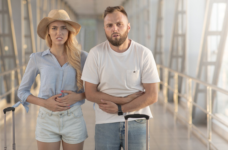 A couple standing in an airport terminal looks sick;  A man with a beard has his hands on his stomach and a woman with long hair has her hands on her stomach.