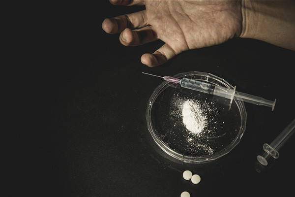 photo representing addiction and overdose: cropped shot of the hand of a person of color positioned next to a small dish with crushed pills and a syringe, with a few whole pills next to it
