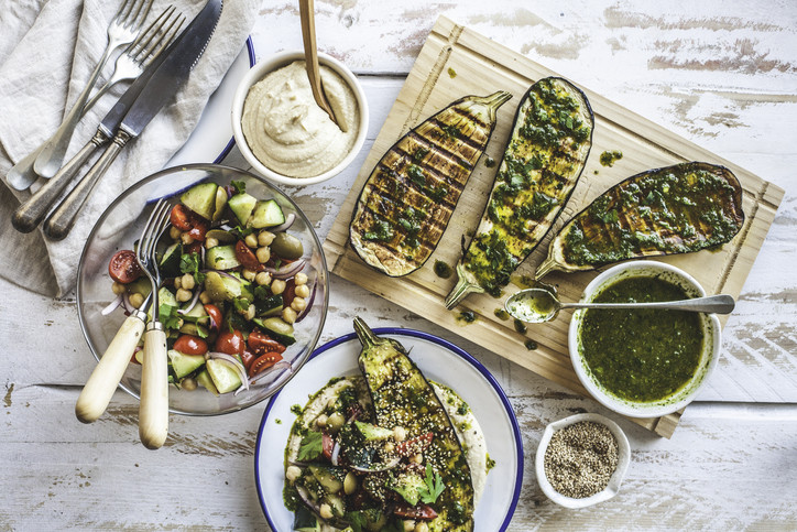Grilled eggplant with a green sauce sits near bowls with hummus, sesame seeds, and a salad with colorful vegetables and beans; 