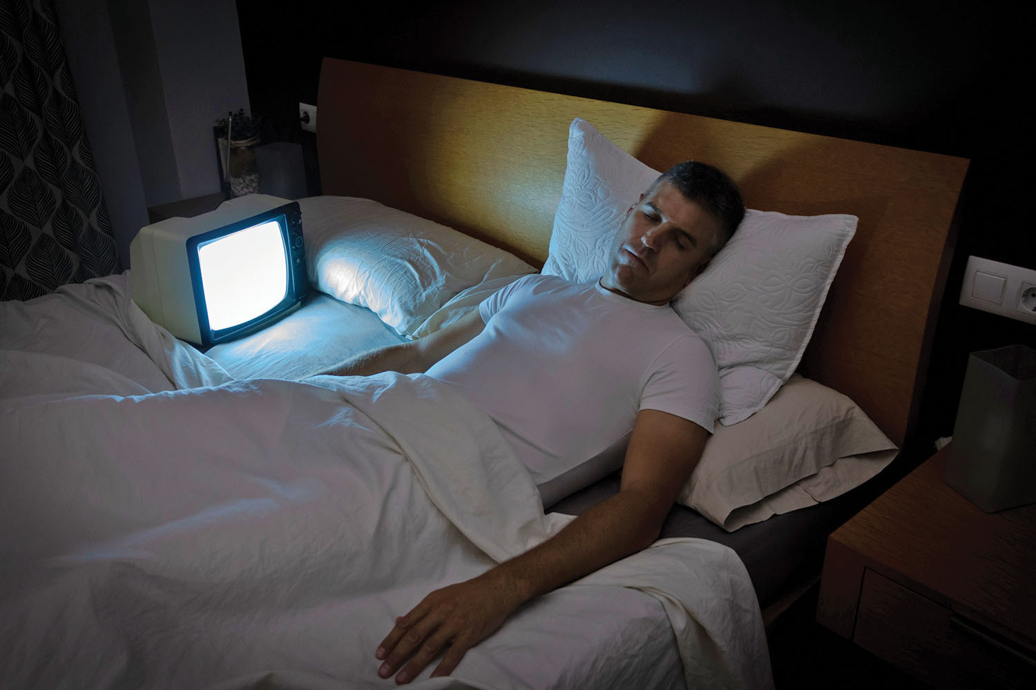 photo of a man asleep in bed with a small TV positioned next to him, throwing light on him