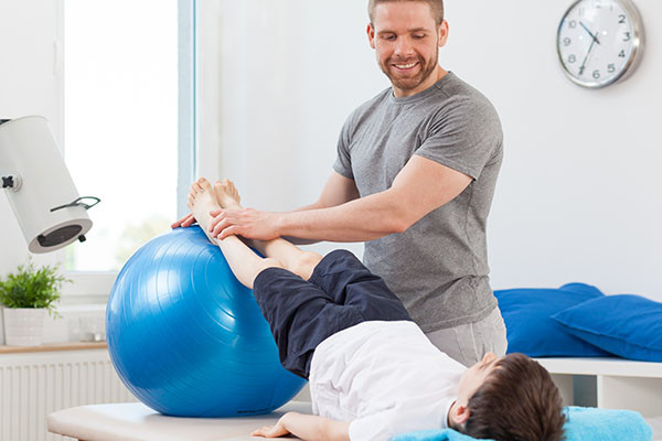 photo of a physical therapist working with a boy who is lying down, with the boy's legs positioned on a balance ball