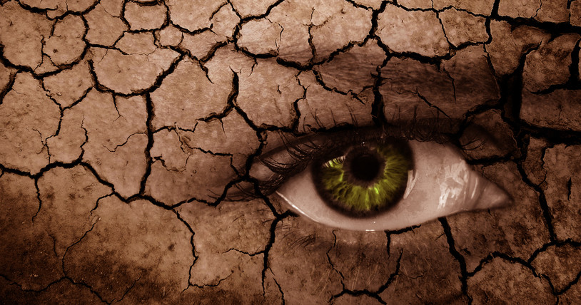 A green eye with a reddened white of the eye looking out from a drought-cracked landscape; concept is dry eye