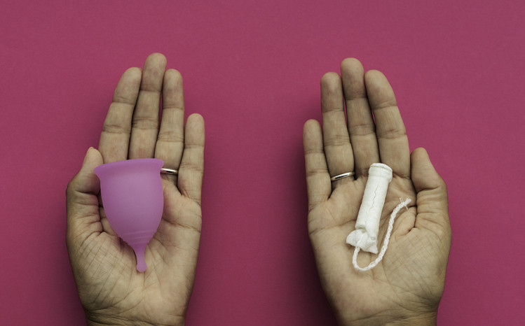 Duas mãos viradas para cima segurando um tampão descartável e um copo menstrual reutilizável, duas opções em produtos menstruais, contra um fundo rosa escuro
