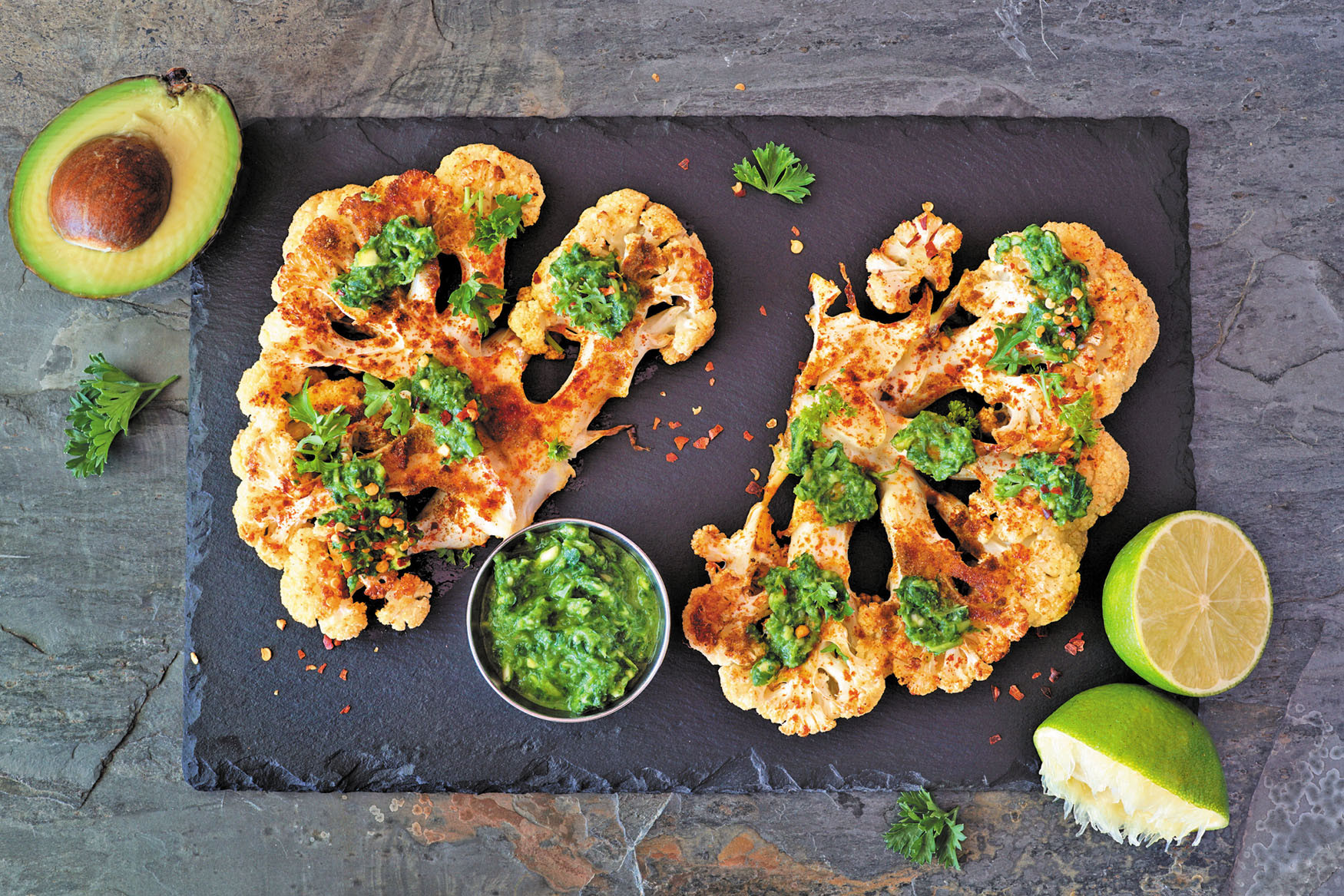 overhead view photo of grilled cauliflower ready for serving, sprinkled with herbs and surrounded by garnishes of avocado, lemon, and lime