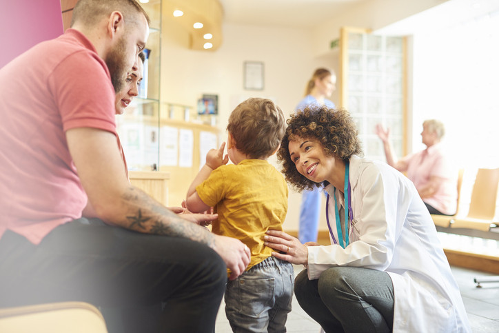 Two seated parents looking on as a pediatrician engages a standing toddler seen from the back in the waiting room of a sunlit health facility