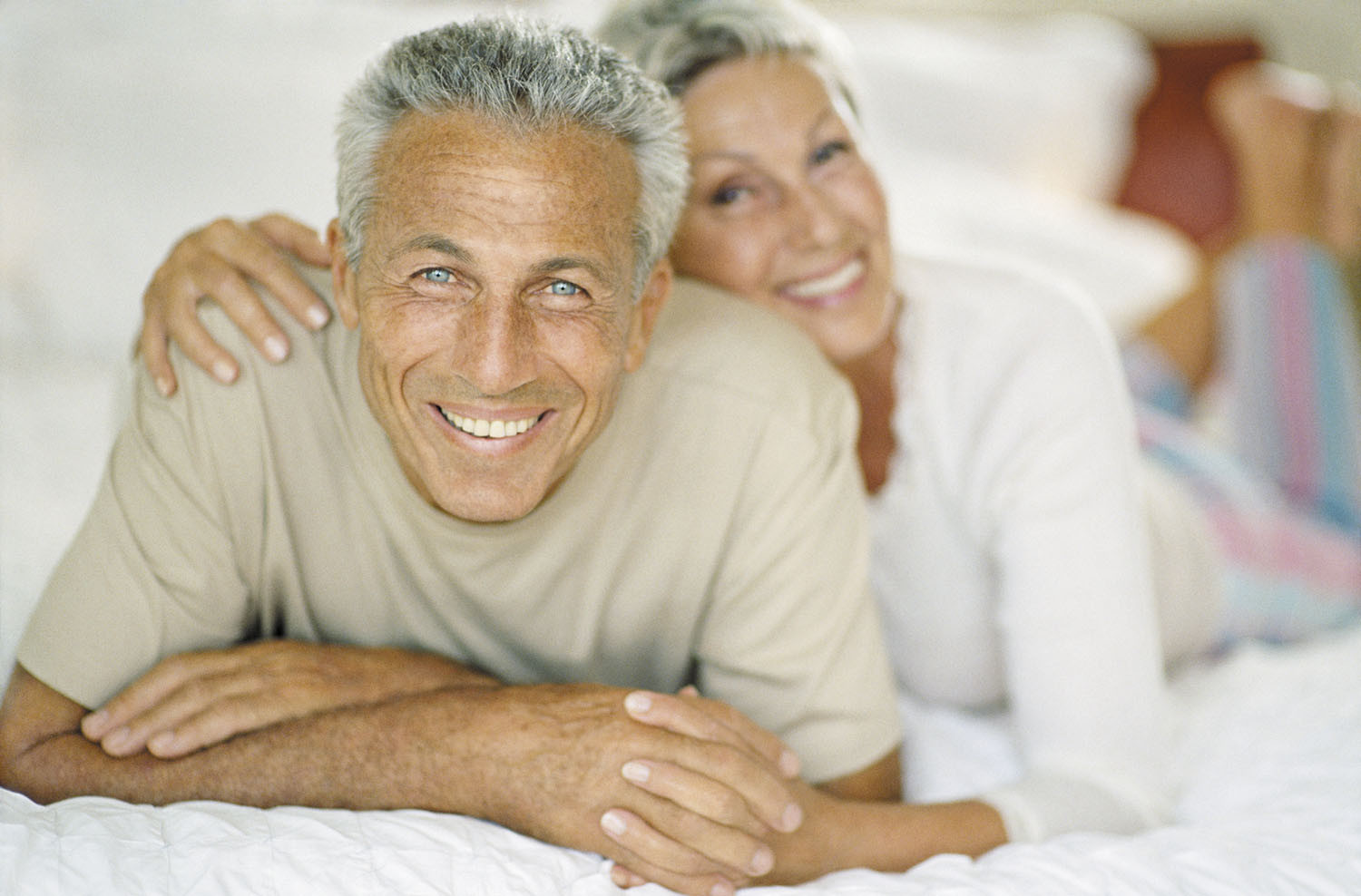 photo of a happy mature couple lounging on their bed, looking at the camera and smiling