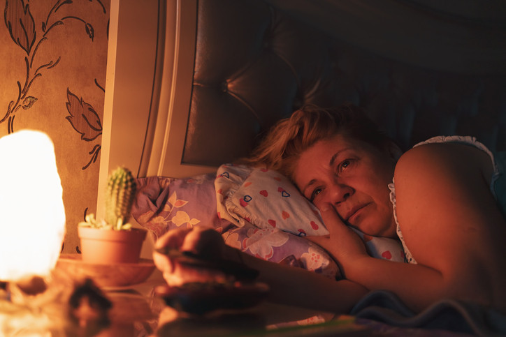 Femme plus âgée allongée sur des oreillers au lit éveillée ;  lumière tamisée et cactus sur la table de chevet, papier peint fleuri en arrière-plan
