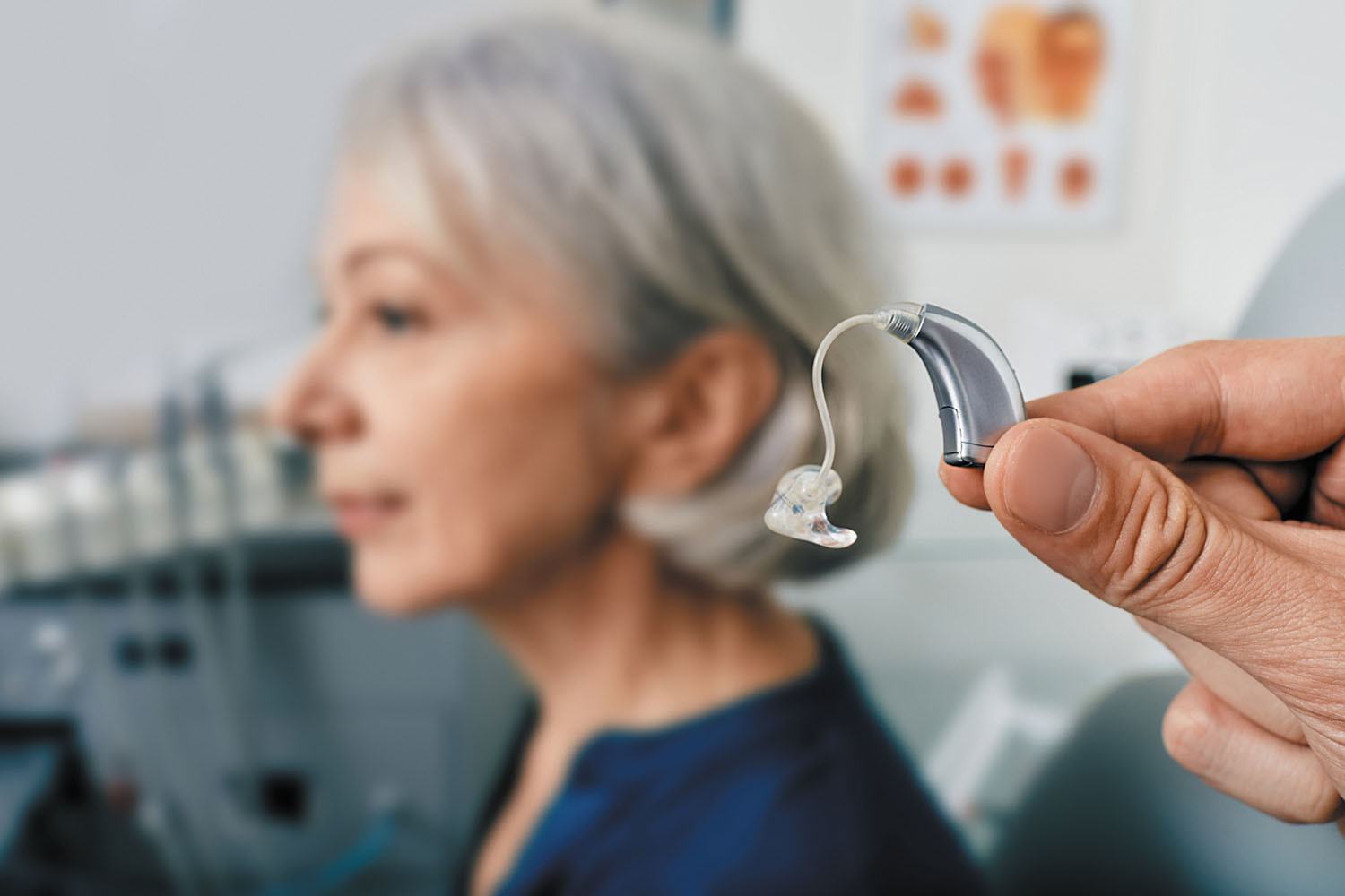 photo of a hand holding a hearing aid in front of a woman slightly out of focus in the background