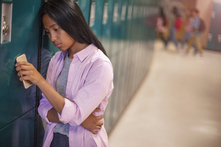An upset middle schooler with her head against the lockers with her phone in her hand