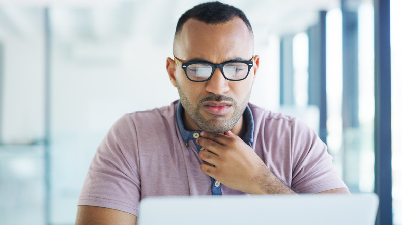 Man in front of lap top at office with uncomfortable look on his face as he tries to clear his throat; he is touching his throat with one hand