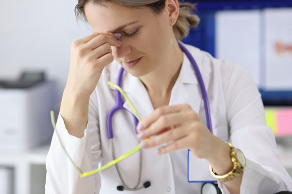 photo of a tired doctor holding the bridge of her nose with her right hand, her safety glasses are in her left hand