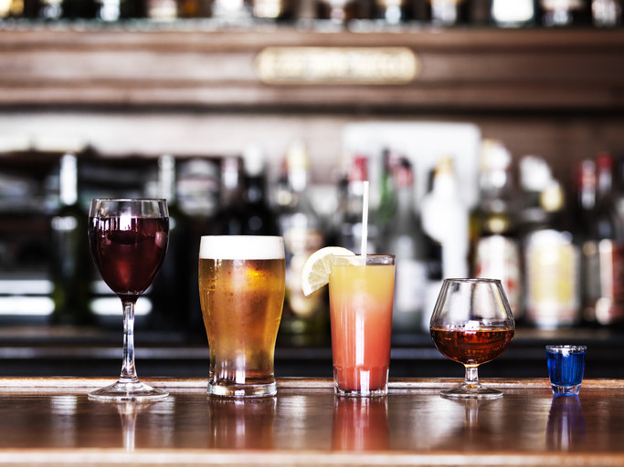 Assorted alcoholic drinks (wine, beer, cocktail, brandy, and shot of liquor) lined up on dark wood bar; blurred alcohol bottles in background