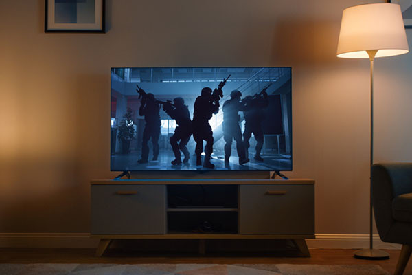 photo of a TV showing a group of armed soldiers in tactical gear moving through an indoor space; TV is in a living room with a lit floor lamp to its right