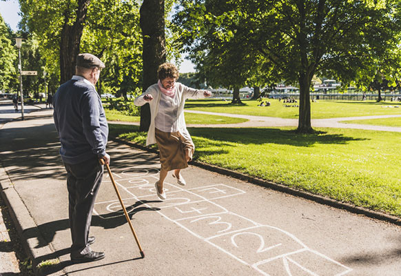 photo of a senior woman playing hopscotch on a sidewalk as her husband watches her