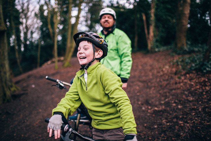 Father behind laughing son, both on bikes on a trail with trees behind them