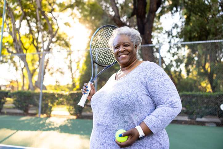 Older woman in lilac top on a tennis court in a park, holding a tennis racket in one hand and a tennis ball in the other