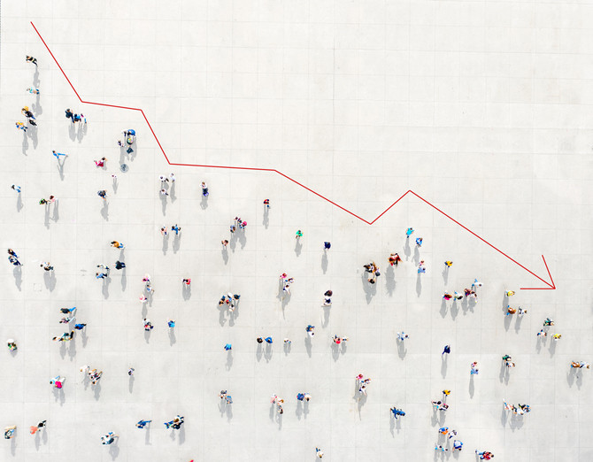 Colorful crowd of tiny people viewed from above hemmed in by a red chart line moving downward 