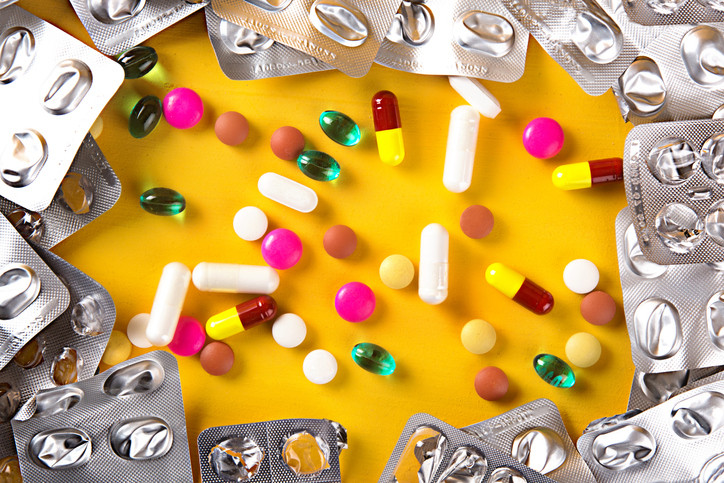 Multicolored pills, tablets, and gel medicines spilling onto a bright yellow background and surrounded by emptied silver blister packs for medications