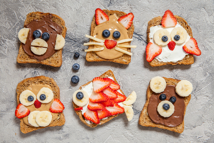 Six pieces of whole wheat toast decorated with fun animal faces added using nut butter, cheese, a chocolatey spread, berries and banana slices