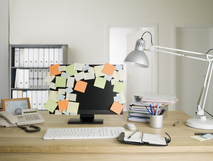 A desk with pens, phone, keyboard, lamp, and computer monitor cluttered with sticky notes 