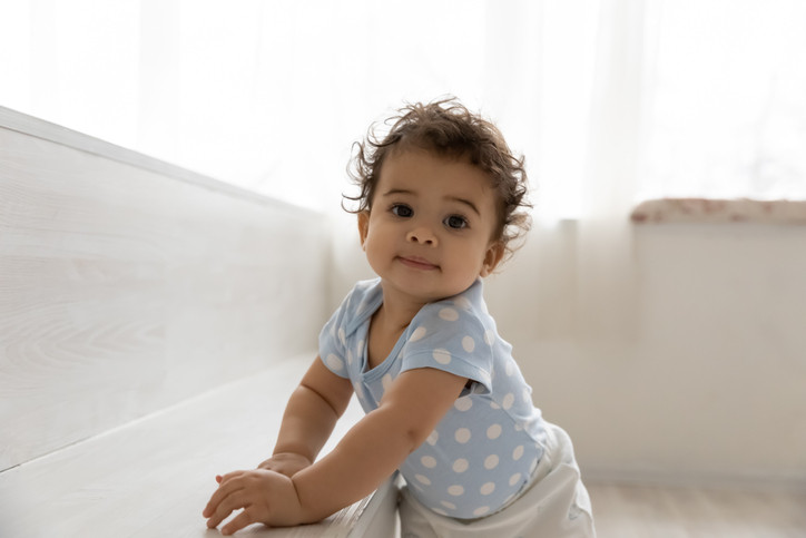 Baby with brown eyes and curly brown hair wearing a polka-dotted blue shirt and holding onto furniture to walk