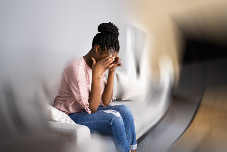 A woman sitting on a sofa, she is rubbing her temples, the room around her is blurry and spinning