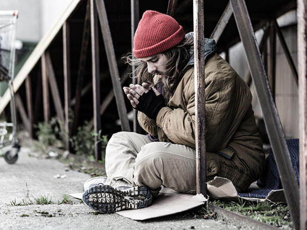 A homeless man sitting cross-legged on a piece of cardboard on the ground, his eyes are closed, and hands are held together