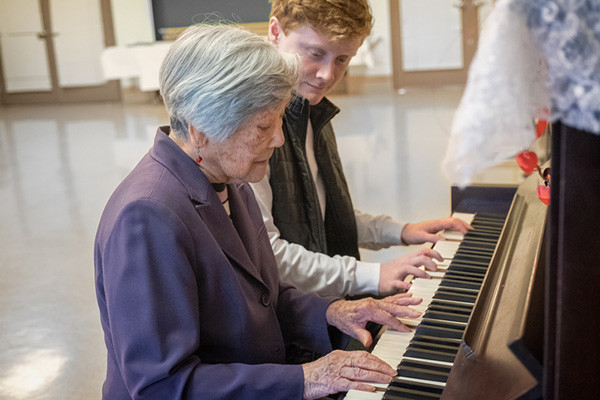 photo of a music therapy session: senior woman is playing piano and a young man is sitting on the bench next to her