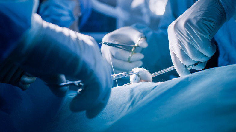 Close up of a surgeon's gloved hands suturing a patient's incision during an operation. Other members of surgical team in background.