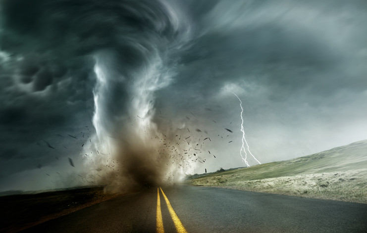 A powerful, destructive storm producing a tornado crosses through fields and roads, throwing debris up into the air as lightening forks down in the distance