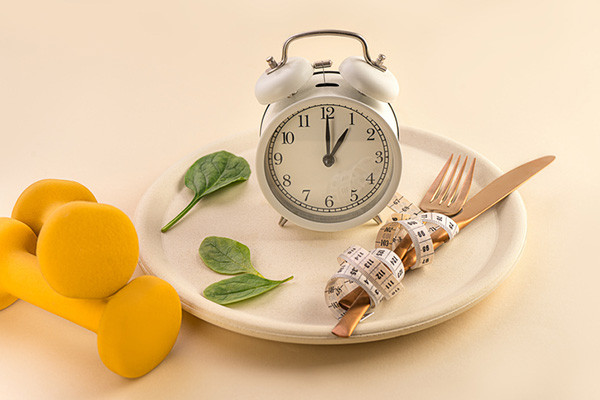 photo of a plate with an alarm clock on it, silverware wrapped in a measuring tape, and a few salad green leaves; next to the plate is a pair of yellow hand weights