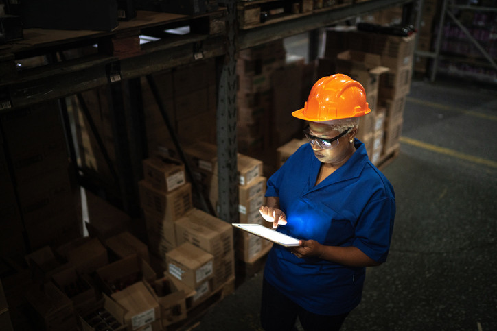Woman wearing blue uniform and orange hardhat standing in aisle of darkened warehouse full of packages typing on lit-up tablet; concept is late shift work