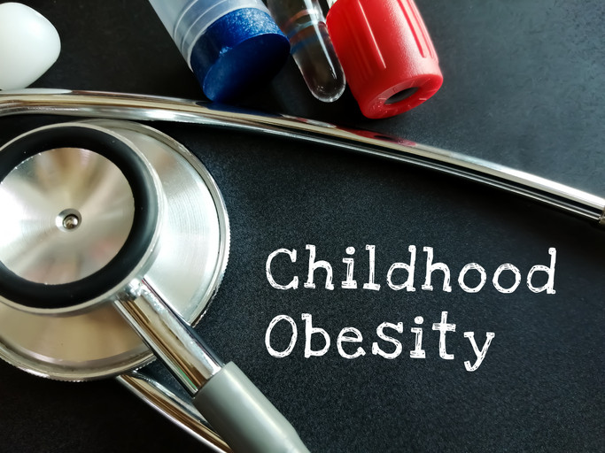 The silver bell of a stethoscope, red and blue tops of testing tubes, and the words "Childhood Obesity" against a dark background