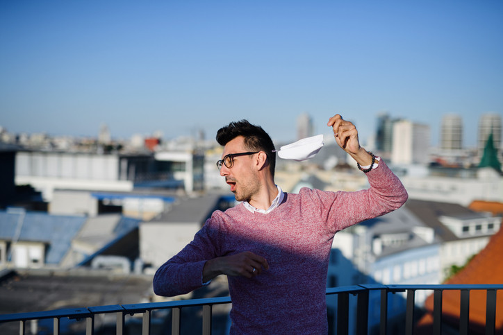 Man with dark hair and glasses standing outside near a railing with a cityscape in the background and ripping off his medical mask