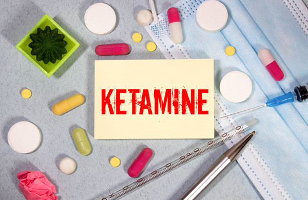 overhead view photo of a yellow post-it pad stamped with the word ketamine in red, surrounded by a pen, a syringe, and an assortment of pills