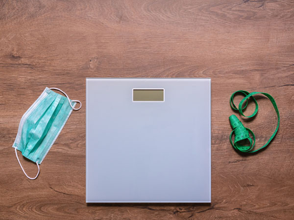 Three objects on a wooden floor, on the left is a disposable face mask, in the middle is an electronic body scale, and on the right is a tape measure